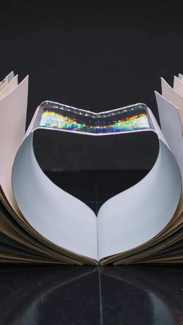Video verticale: Dare forma alle pagine di un libro da cerchio a cuore, poi aprirle a ventaglio su un tavolo lucido, con una striscia olografica visibile.