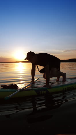 man wearing sunglasses, cap, t-shirt and shorts sits on the sup board floating on water. sportsman stands up and rows away. low angle view. vertical video.