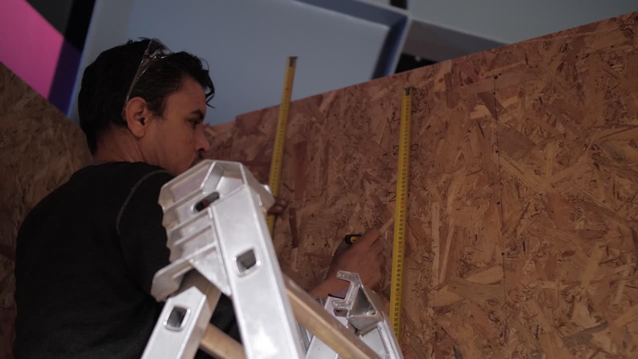 Worker measuring wall height with a tape measure while standing on a ladder