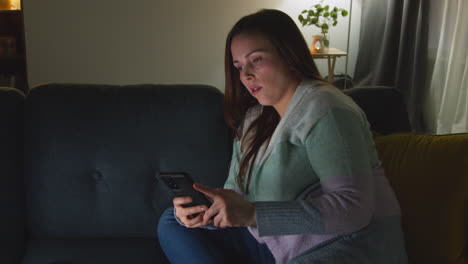 woman sitting on sofa at home at night streaming or watching movie or show on laptop and scrolling internet on phone 10