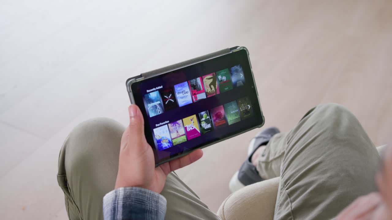 Uomo afroamericano che tiene un tablet, con la mano destra tocca e scorre i contenuti multimediali mentre naviga in salotto.