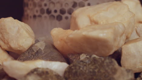 close up of steaming sauna stones glowing on metal heater grate against warm wood panels with rising vapor, textured rock detail in soft light, creating serene wellness ritual atmosphere