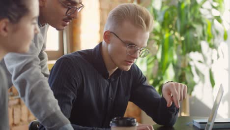 coworkers talking about a project, one is sitting on desk in front of the computer and the others are standing