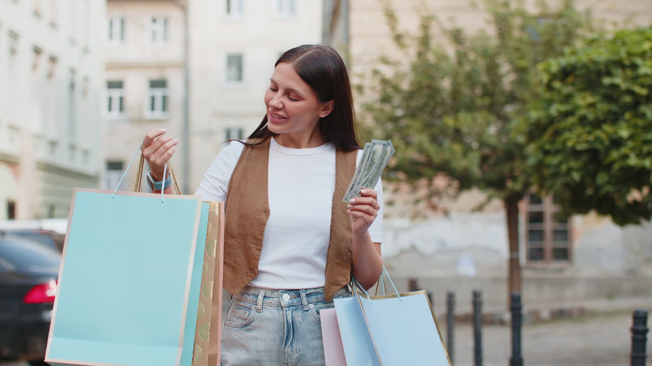 Happy young shopaholic caucasian woman holding cash while carrying shopping bags on city street