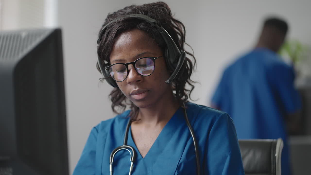 A black woman doctor wearing headphones sits at a table with a computer and takes calls from patients looks at their medical records and enters them into the clinic schedule