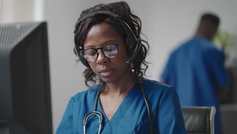 A-black-woman-doctor-wearing-headphones-sits-at-a-table-with-a-computer-and-takes-calls-from-patients-looks-at-their-medical-records-and-enters-them-into-the-clinic-schedule