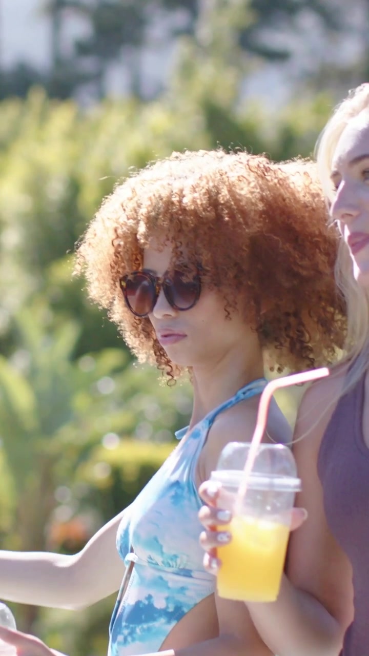 Vertical video: Receiving curly woman accepting cup after handoff at poolside, in swimsuit toasting