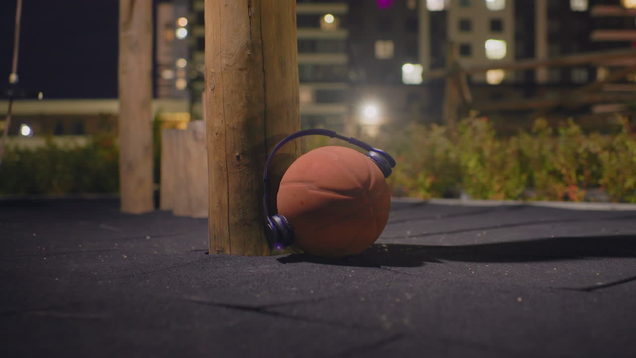 Partita di basket notturna, appoggiato a un palo di legno, cuffie appoggiate sulla palla, superficie di asfalto crepata, luci della città in lontananza, bokeh dello skyline sfocato, panchina del parco giochi vuota, tenue luce del lampione, cinematografico