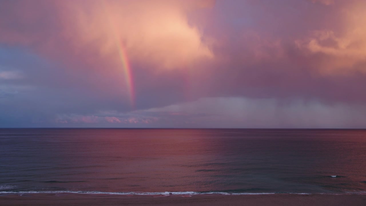 嵐の後の夕暮れ時、穏やかな海にかかる虹。海面に映る劇的な雲、色鮮やかな海岸の風景、そして幻想的な気象現象