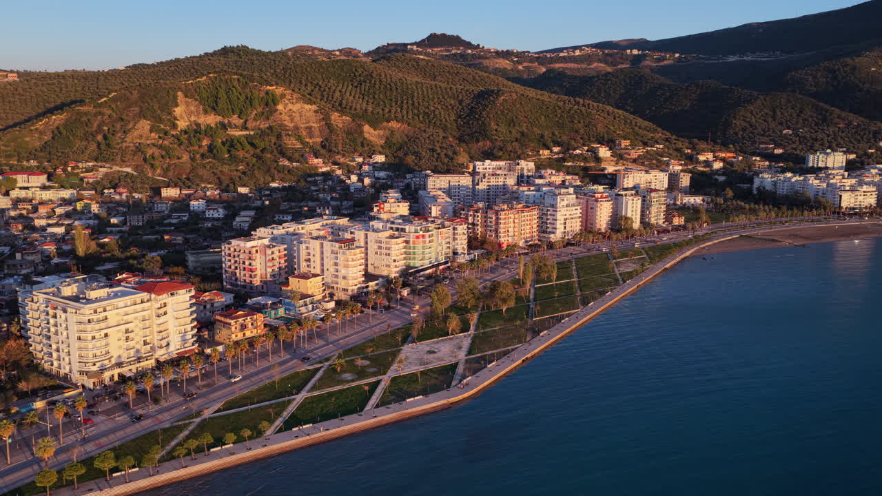 Aerial drone view of Vlora's modern coastline at golden hour, with warm sunlight illuminating residential buildings and lush forested hills in the background