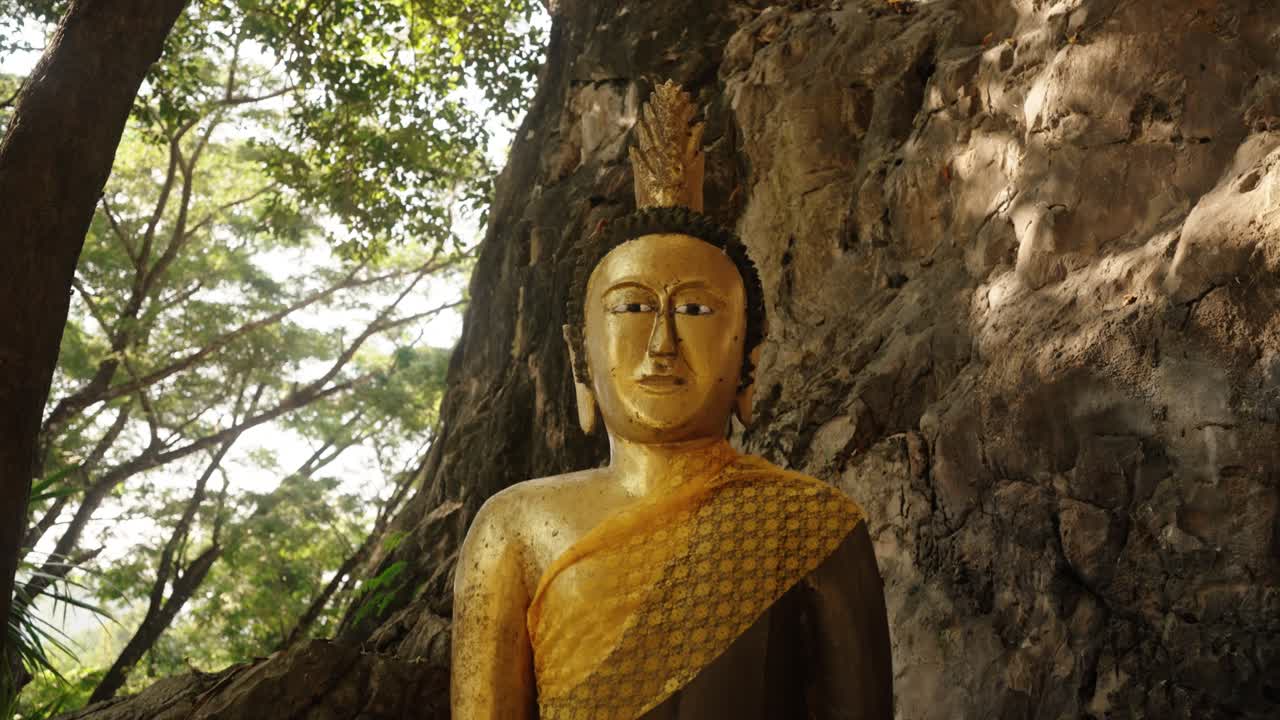 A golden seated Buddha rests peacefully beside a rocky cliff at Wat Tham Phra