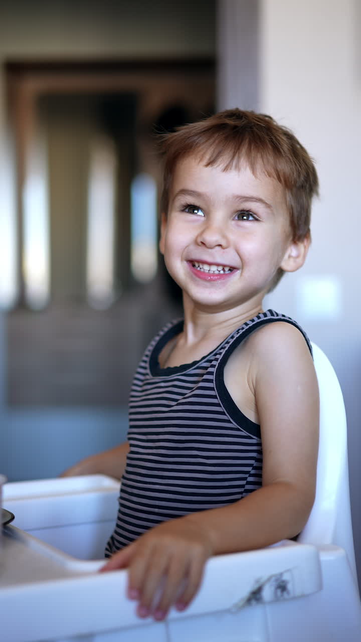 Cute smiling Caucasian kid sits at high chair. Mom's hand passes a full spoon to a baby but he refuses to eat. Vertical video.