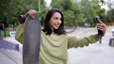 Portrait-of-a-brunette-in-a-green-sweater,-who-sits-on-the-background-of-the-park-with-a-skateboard-in-her-hands-and-takes-a