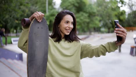 ritratto di una bruna in un maglione verde, che si siede sullo sfondo del parco con uno skateboard nelle sue mani e prende un