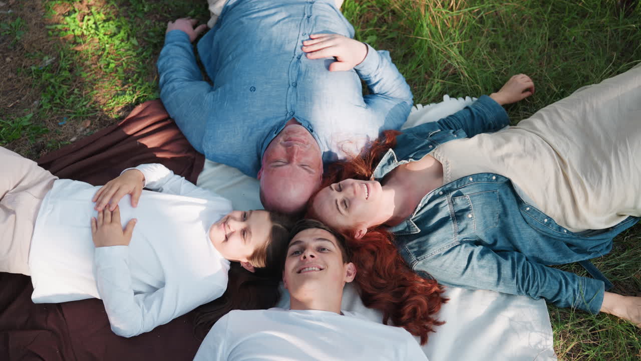Aerial view of outdoor family lying down on blanket with heads joined as son talks and others listen, enjoying warm sunlight, peaceful atmosphere, and emotional bond during relaxing picnic moment