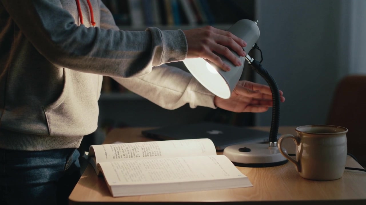 Ein Student im Kapuzenpullover greift nach der Lampe, stellt die Schreibtischlampe ein und beleuchtet ein offenes Buch am Schreibtisch zum Lesen.