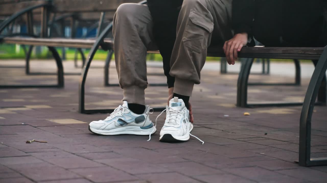 Person binder hvide sneakers på en bænk, nærbillede af hænder, der binder snørebånd, hvide sportssko på tekstureret belægning, cargobukser, metalbænkestel, byparkstemning, blødt morgenlys, flygtigt.