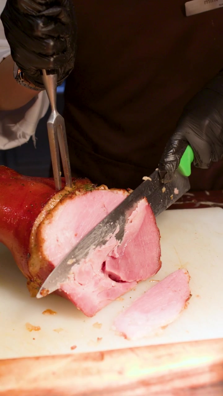 Uno chef professionista con guanti neri affetta prosciutto arrosto su un tagliere bianco.