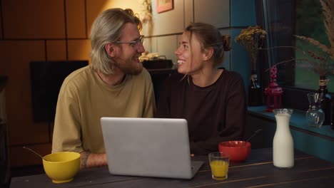 feliz chico rubio con barba en gafas y una chica rubia se divierten y miran la pantalla de la computadora portátil por la noche en una habitación iluminada por una lámpara amarilla