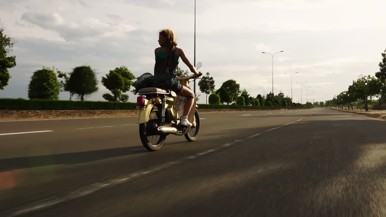 The beautiful young blonde caucasian girl is riding on an empty highway on a yellow scooter - dolly shot from behind
