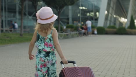 child girl tourist with suitcase near airport. kid wave hand, walks down street with bag. vacation