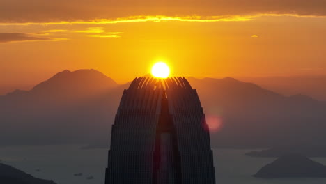 Toma-Aérea-Ascendente-Que-Muestra-La-Silueta-Del-Edificio-De-La-Ifc-Al-Atardecer-Dorado-En-Hong-Kong---Toma-De-Zoom-Con-La-Cordillera-De-Fondo