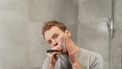 handsome young man shaving in front of a mirror with a dangerous razor