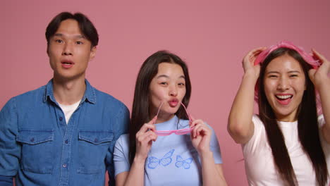 핑크색 스튜디오 배경 앞에 있는 젊은 친구 그룹이 장비로 사진 부스 스타일의 초상화를 위해 포즈를 취하고 있습니다.