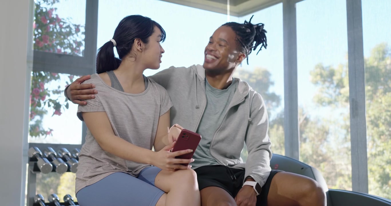 Fit couple sitting on bench in sportswear woman tapping red phone sharing as man pointing laughing