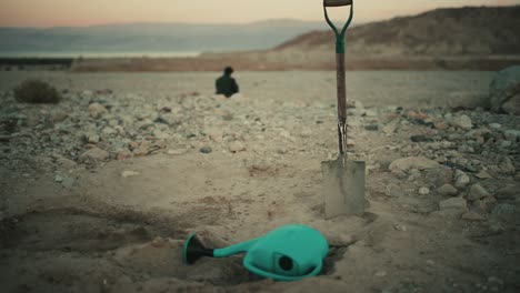 Watering-can-and-shovel-on-sand-with-nomad-man-silhouette-in-background-at-sunset