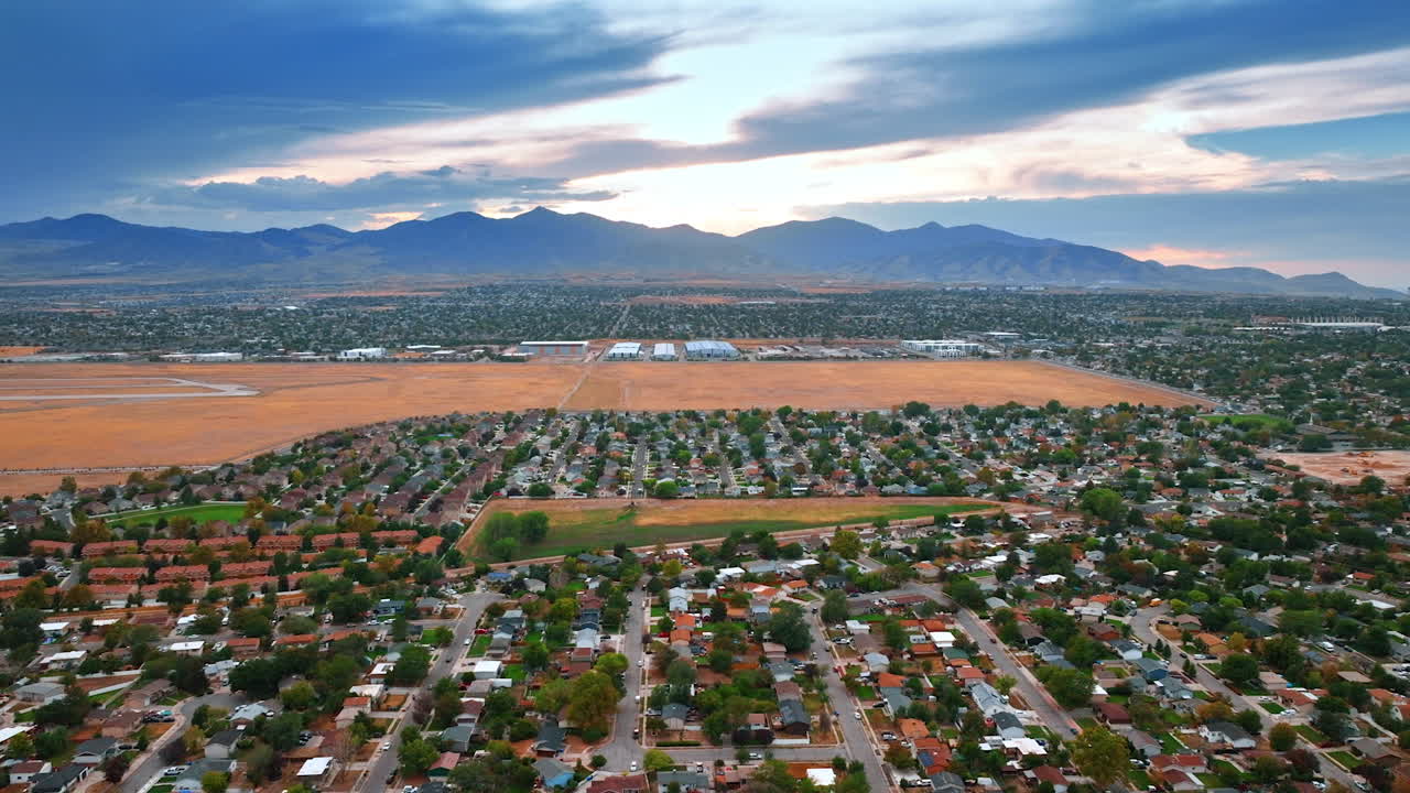 Green low-rise residential area with a bare field in center. Flight above the scenery of Salt Lake City, Utah, USA at sunset. Mountain range silhouette at backdrop