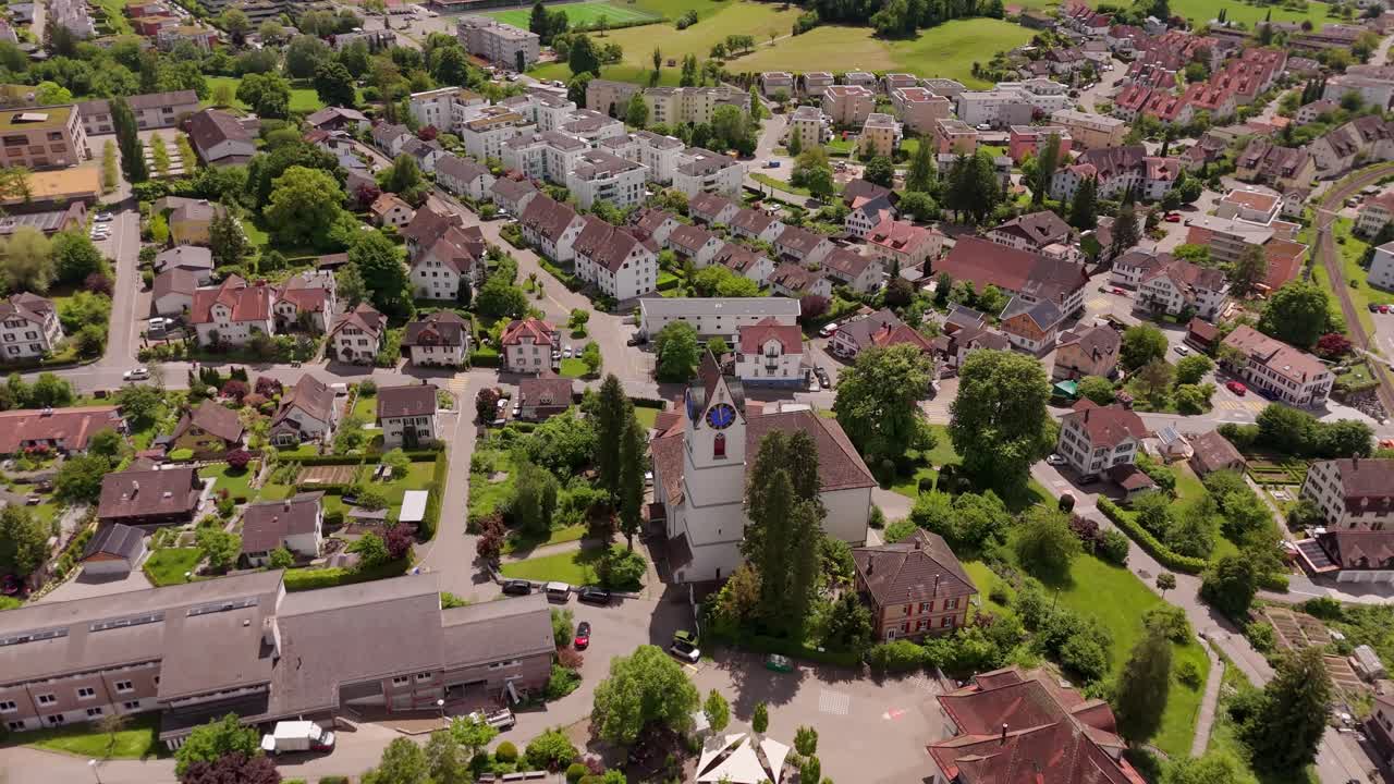 Historic church tower in Hinwil a swiss town in summer. Aerial top down orbit shot. Red roofs of houses and luxury apartment Blocks in Switzerland. Green grass and trees.