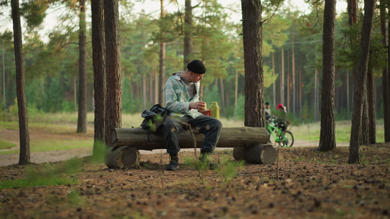 Kaukasisk vandrer hviler sig på en træstamme i en stille fyrreskov, tjekker kort og pakker en snack ud af rygsækken ved siden af sig, drikker vand og spejder efter stien, mens en cyklist kører forbi i baggrunden. Blødt sollys filtreres.