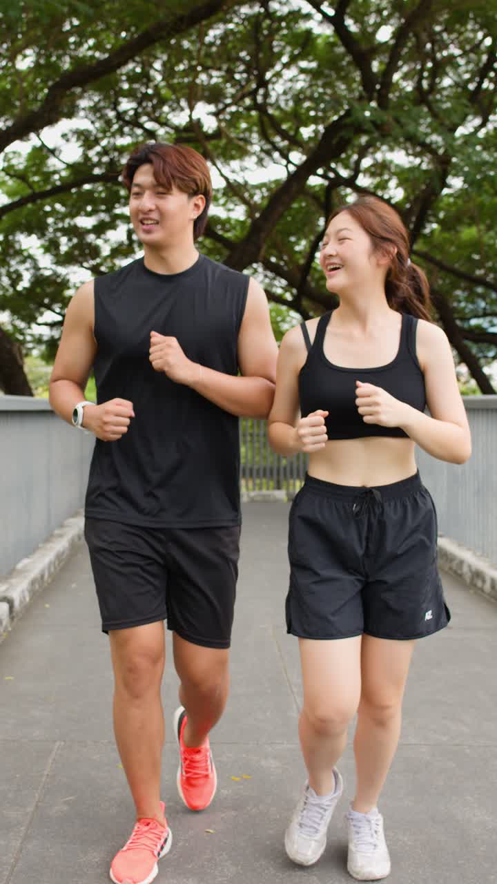 Coppia asiatica sorridente fa jogging fianco a fianco lungo un sentiero alberato di un parco urbano, ripresa a mano libera con movimento in avanti.