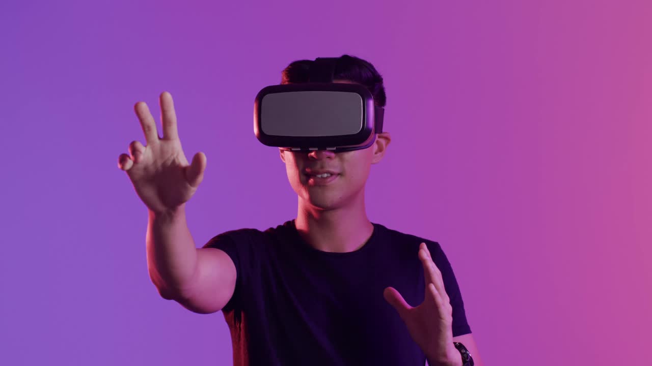 Happy asian man using vr headset and touching virtual screen on purple background, slow motion