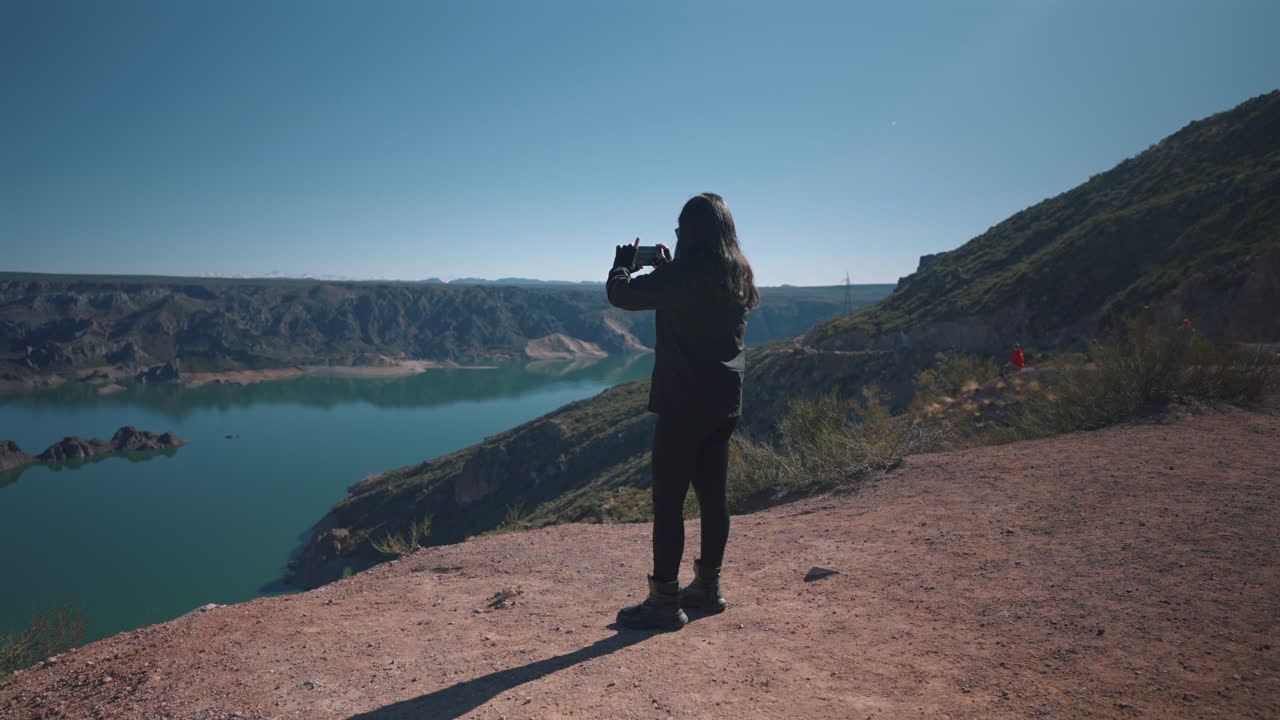 Static slow shot frames girl holding smartphone shooting photo toward turquoise reservoir and canyon rims from dusty overlook in Cañón del Atuel, visitor engagement with Mendoza's valley vista