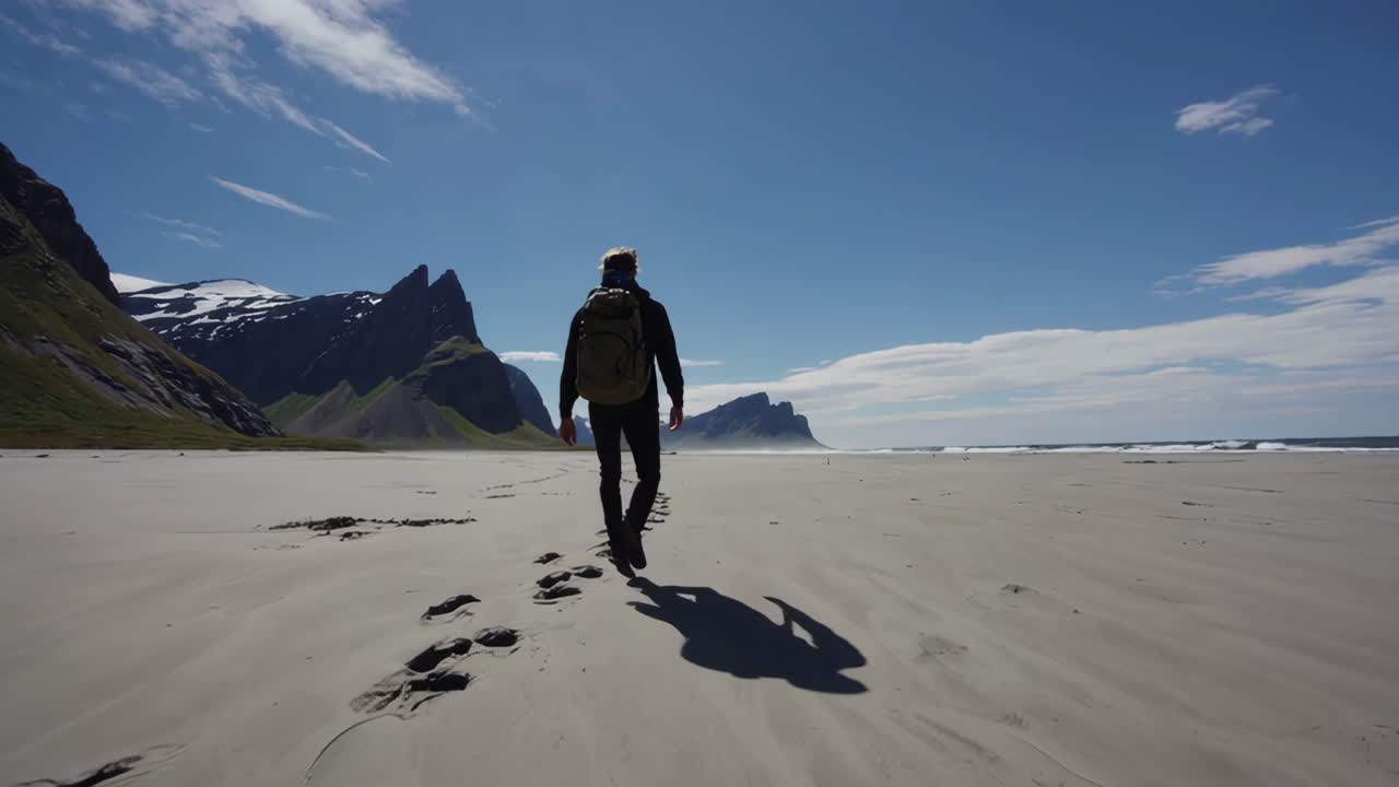 A person walks along a vast sandy beach towards majestic mountains under a blue sky