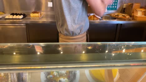 Young-man-standing-at-the-bakery-counter-and-selling-bread-or-cakes-as-he-prepares-it-with-several-brigadeiros-behind-him