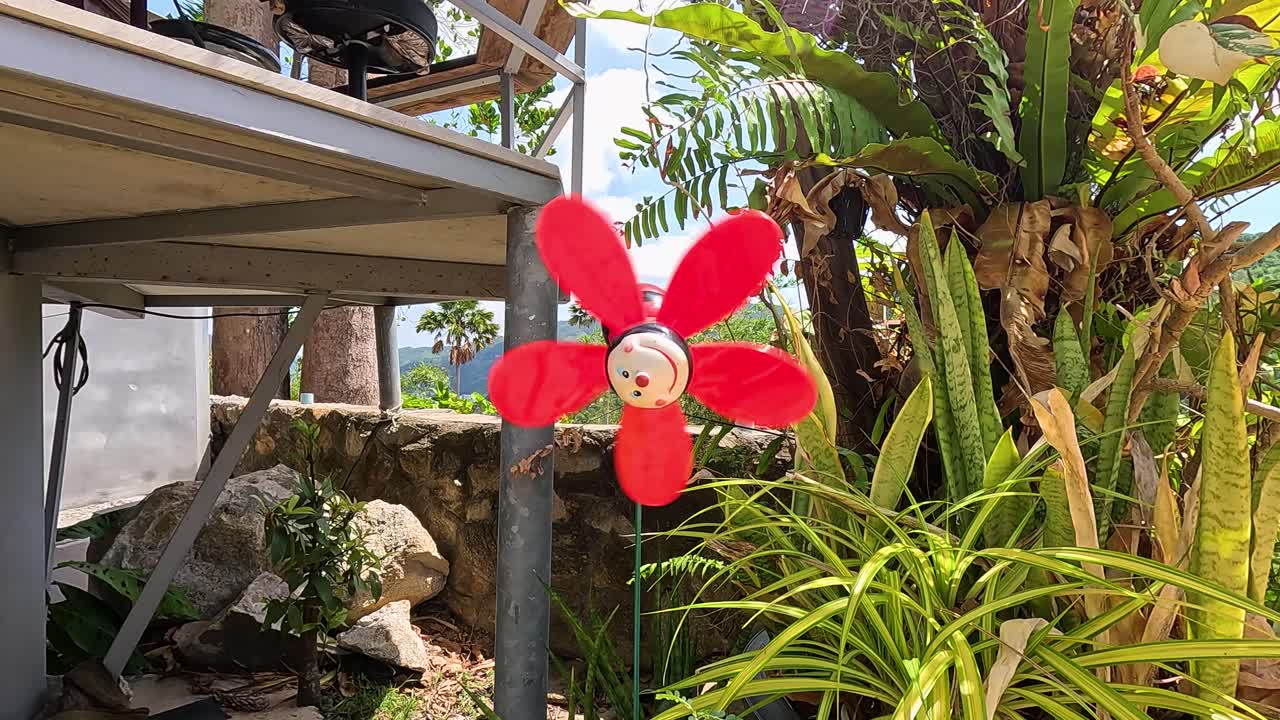 A vibrant red flower windmill spins in a lush tropical garden in Phuket, Thailand, under bright daylight