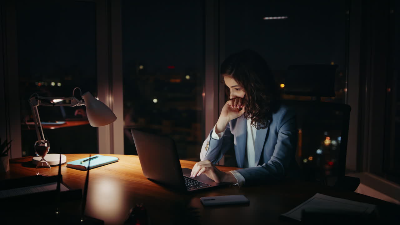 Empresaria somnolienta trabajando de noche sentada en el escritorio de la oficina, mirando su portátil.