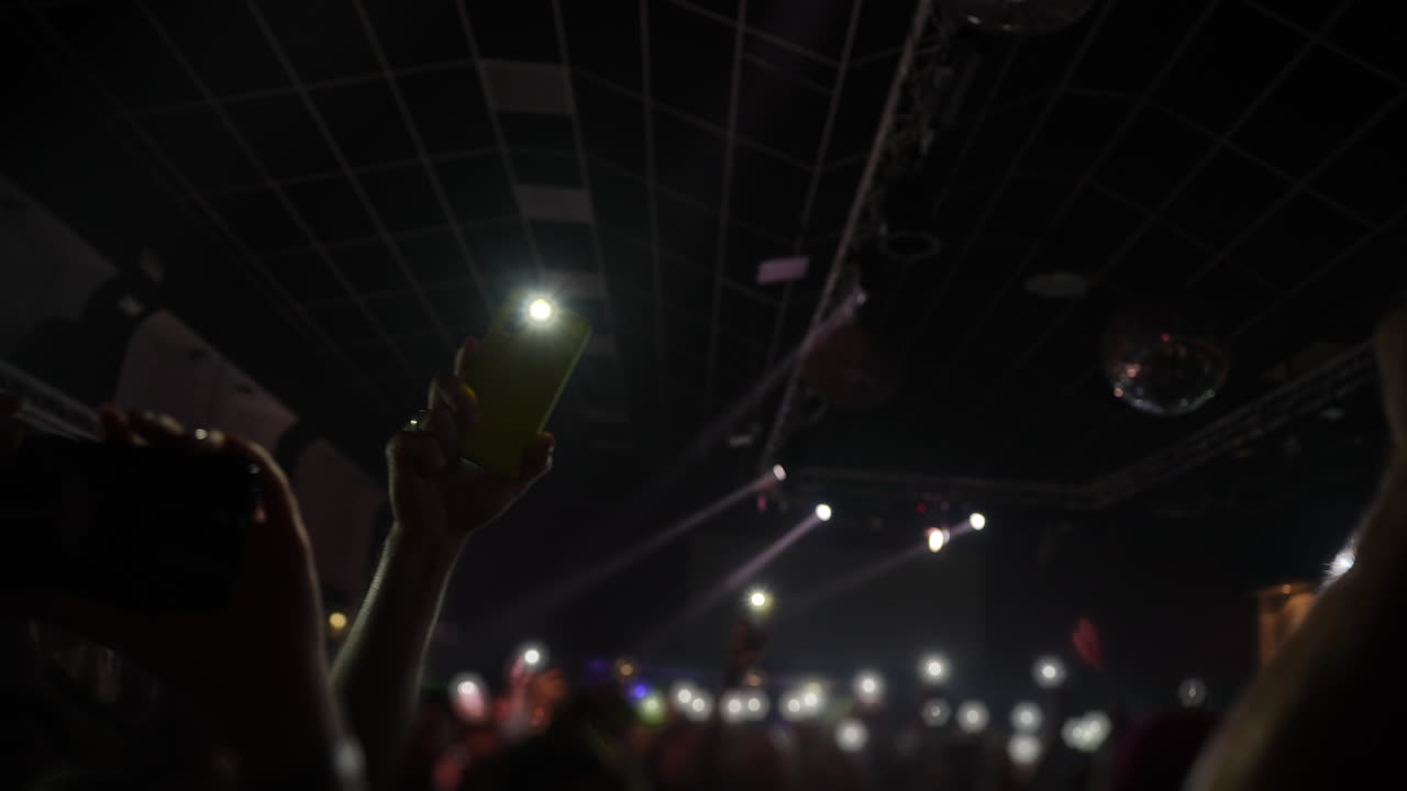 Crowd at concert with raised hands holding smartphones capturing vibrant stage lights