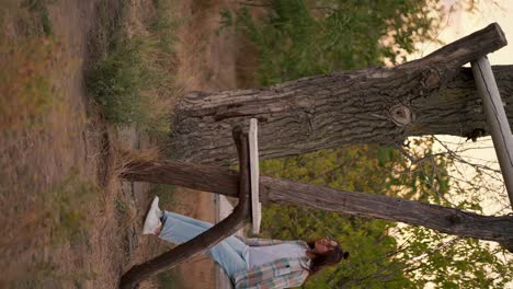 Vertical-video:-a-brunette-in-a-checkered-shirt-and-jeans-swings-on-a-wooden-swing-outside-the-city-against-a-forest-background.-Rest-in-the-country