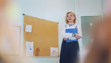 teacher is talking in the class. attractive teacher holding a blue folder telling something to the kids. female educator stands near the pinboard in the classroom background.