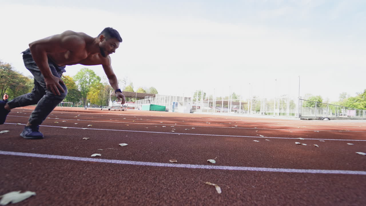 Sportive runner on outdoor athletics track. Athletic man without shirt running on stadium track on green trees background. Motion camera around.