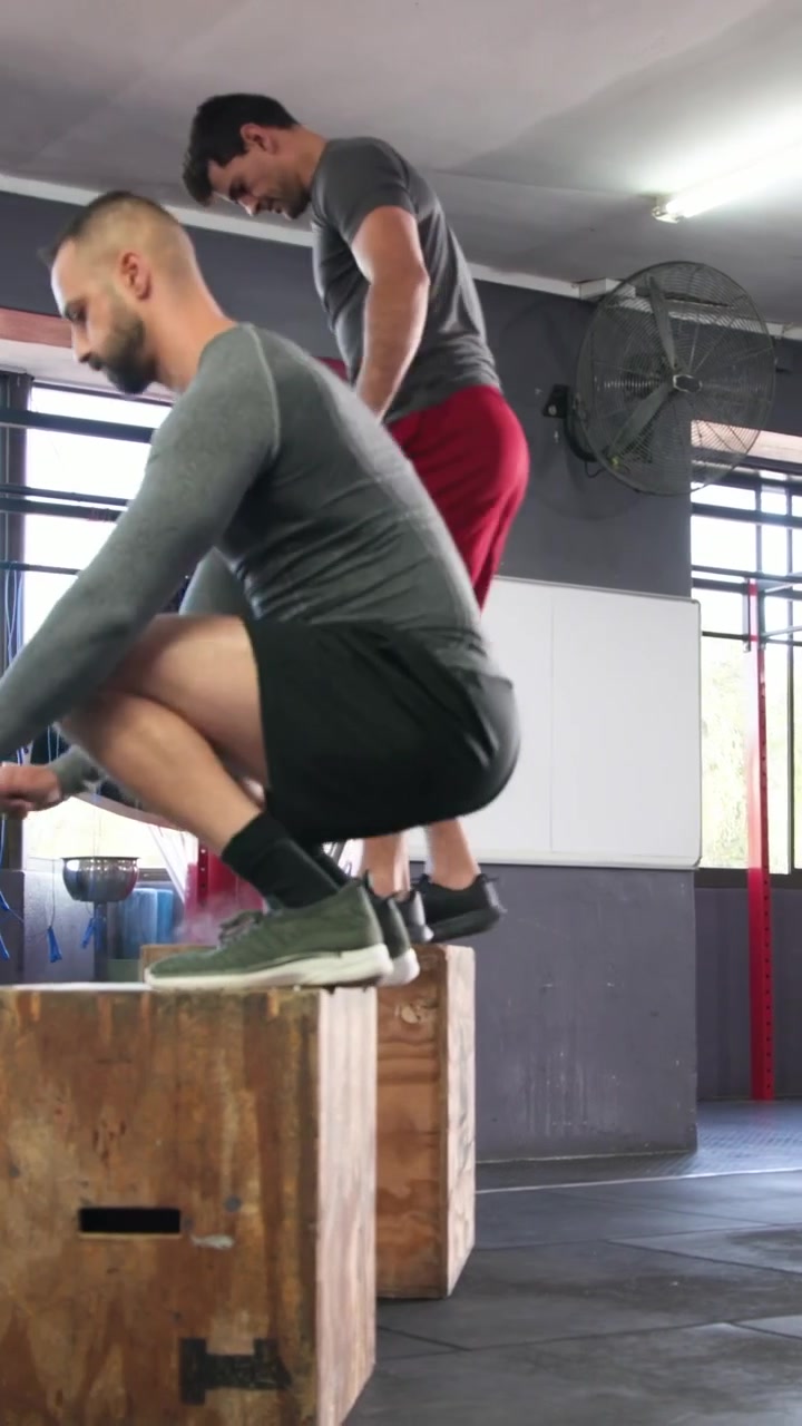 Video vertical: Hombre saltando de espaldas al inicio, dúo de gris haciendo saltos pliométricos en cajón para entrenar en el gimnasio
