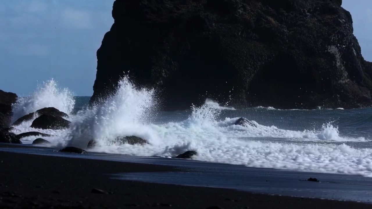 Onde impetuose spinte dalla mareggiata che colpiscono un faraglione e massi su una spiaggia di sabbia nera, sollevando schiuma