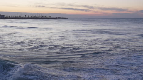 Ola-De-Puesta-De-Sol-Iridiscente-En-La-Costa,-Con-Fondo-De-Muelle-De-Horizonte-De-Luz-De-Puesta-De-Sol-Dorada