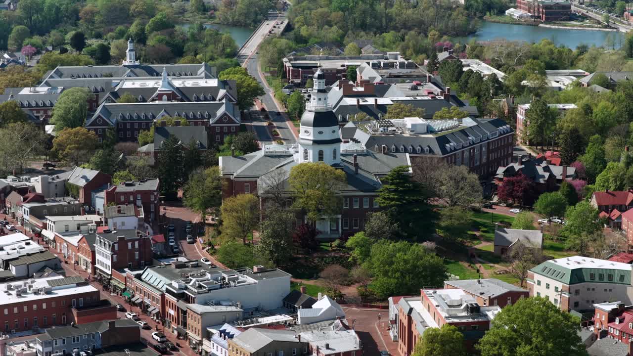 Cảnh quay drone từ trên không Annapolis lịch sử, Maryland, Hoa Kỳ với Tòa nhà Quốc hội Maryland, những con phố cổ, các tòa nhà gạch đỏ, dòng sông êm đềm, cầu và khung cảnh thành phố mùa xuân tươi tốt dưới ánh nắng mặt trời. Cảnh quay xoay quanh