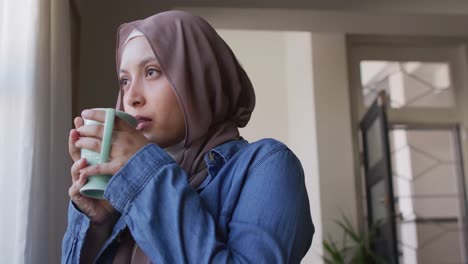 video of happy biracial woman in hijab drinking coffee, looking out of window at home