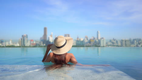 Mujer-Solitaria-Con-Sombrero-Flexible-En-La-Piscina-Infinita-De-La-Azotea-Con-Vistas-Al-Paisaje-Urbano-Soleado,-Vista-Trasera